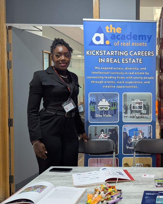 Person standing beside an information table at an indoor event, with a banner reading ‘the academy of real assets – Kickstarting careers in real estate’ behind them. 