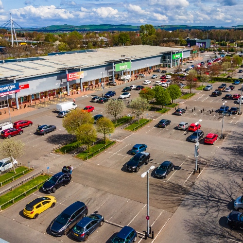 Aerial view of a retail park with a large shopping unit, car park filled with parked vehicles, and people walking between stores along a covered walkway