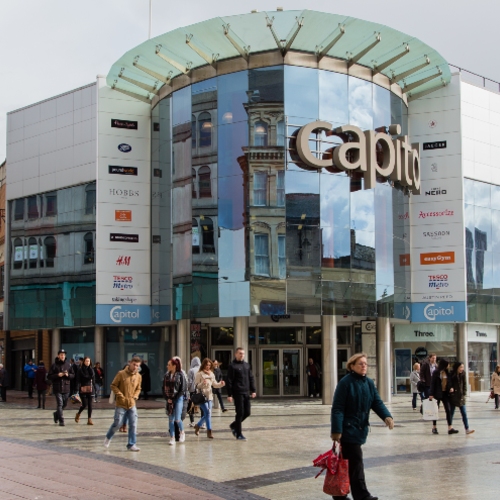 Exterior of a shopping centre with a glass‑fronted entrance displaying ‘Capitol’ signage, surrounded by retail brand logos and people walking across a public plaza in front of the building