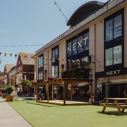 Exterior of a shopping centre with a Next store, featuring an open pedestrian area with seating, planters, and decorative bunting, and people walking and sitting outside