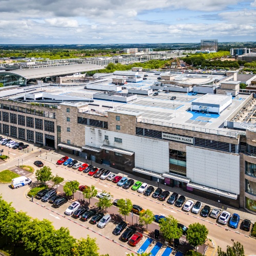 Aerial view of a large shopping centre with ‘Midsummer Place’ signage, showing rooftop structures, surrounding buildings, and a car park with parked vehicles along the street below.