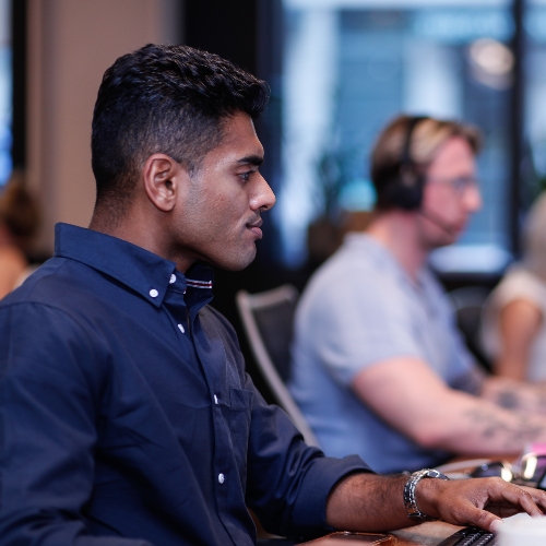 Person seated at a desk using a computer keyboard in an office environment, with other people working at desks in the background, including one person wearing headphones
