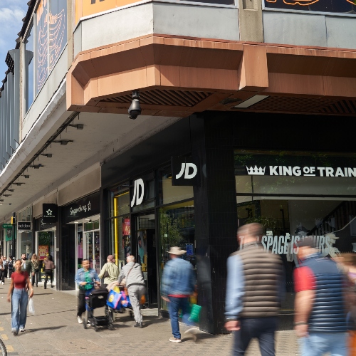 Exterior of a shopping centre high street with retail stores including JD and other shopfronts, and people walking along the pavement outside the entrance